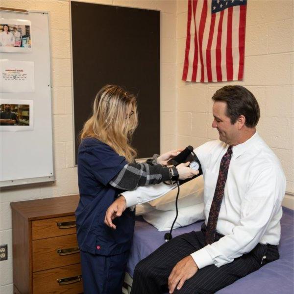 Man in white shirt gets blood pressure check from student in blue scrubs